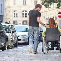 eine Frau in einem Rollstuhl wird von einem Mann begleitet