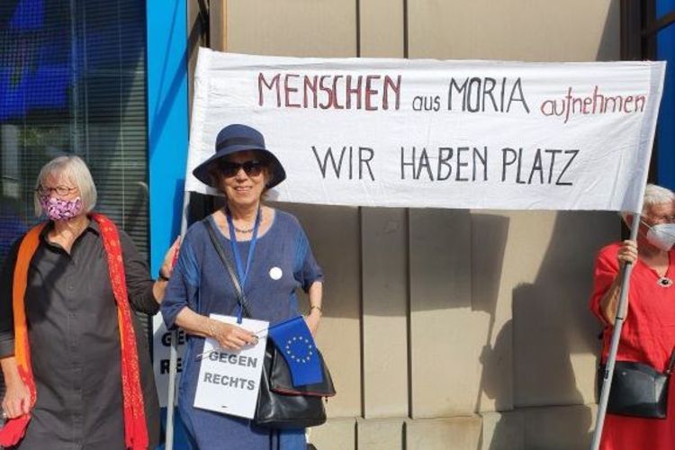 Hilde von Balluseck in einem blauen Kostüm vor einem Transparent mit der Aufschrift: Menschen aus Moria aufnehmen - Wir haben Platz