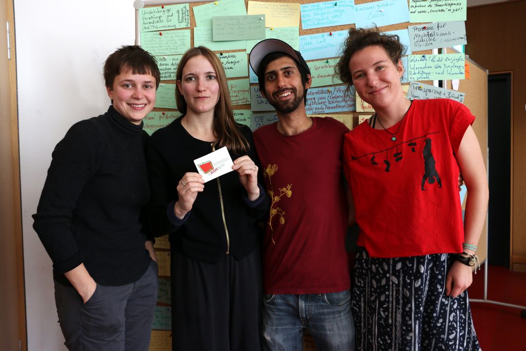 Vergrößern: Vier junge Menschen stehen nebeneinander vor einer Stellwand mit farbigen Karten, auf denen mit Edding etwas notiert wurde, und schauen fröhlich in die Kamera. Die zweite von links hält einen Aufkleber, auf dem ein rotes Viereck mit grünen Pfeilen drumherum abgebildet und das Wort "trASHform" zu lesen ist.