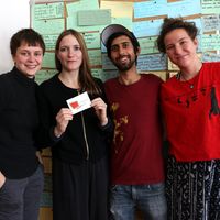 Vier junge Menschen stehen nebeneinander vor einer Stellwand mit farbigen Karten, auf denen mit Edding etwas notiert wurde, und schauen fröhlich in die Kamera. Die zweite von links hält einen Aufkleber, auf dem ein rotes Viereck mit grünen Pfeilen drumherum abgebildet und das Wort "trASHform" zu lesen ist.