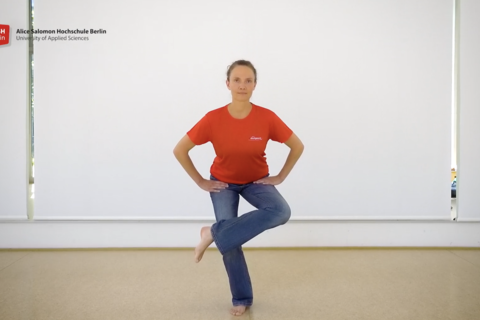 Eine Frau steht im Bewegungsraum der ASH Berlin, die Hände in den Hüften, ein Bein überkreuzt im stehen das andere