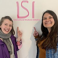 Anja and Chrystal, the two authors and founders in front of a wall pointing at a tag reading "ISI - International Students Initiative"