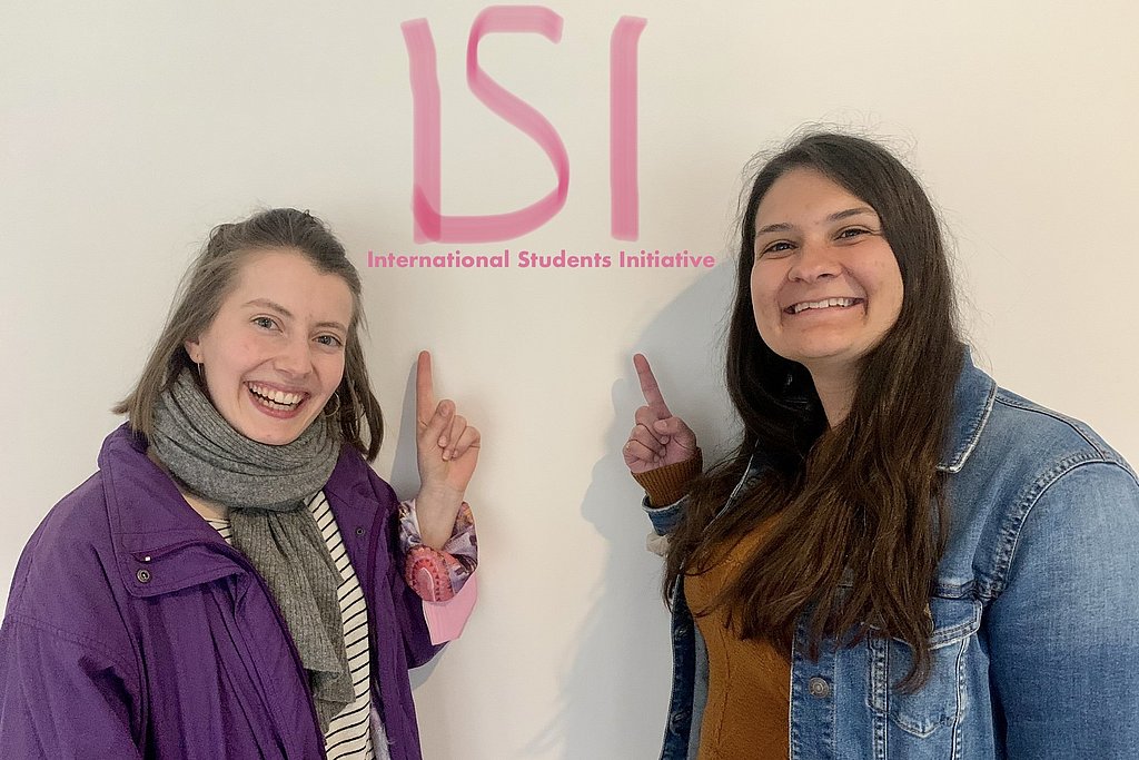 Anja and Chrystal, the two authors and founders in front of a wall pointing at a tag reading "ISI - International Students Initiative"